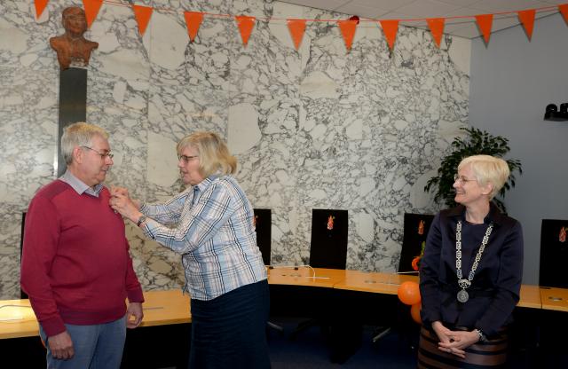 De zus van de heer Van Duin speldt de onderscheiding op, burgemeester Dassen kijkt op afstand toe