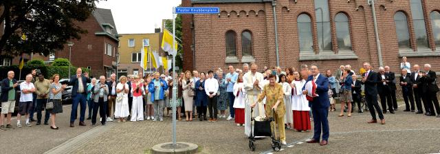 Een grote groep mensen voor de Sint Martinuskerk. Op de voorgrond het straatnaambord, daarnaast staan de wethouder en de zus van Pastor Knubben. 