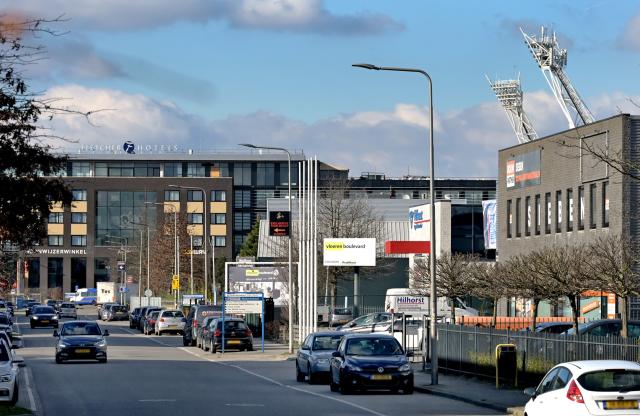 Zicht op het Parkstad Limburgstadion vanuit de Wiebachstraat. Auto's en bedrijfspanden. 