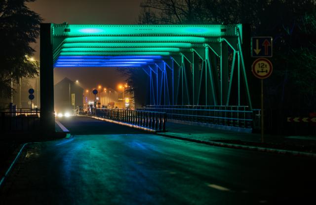 De verlichte spoorbrug in het donker. De brug licht blauw en groen op. Op de achtergrond de rotonde  en de Lidl. 