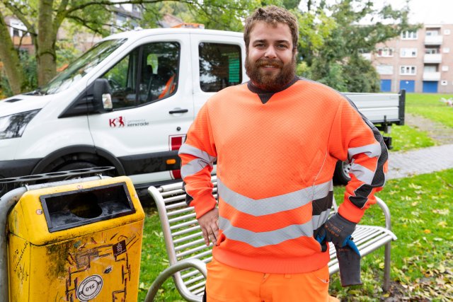 Dennis in zijn oranje werkkleding naast een prullenbak en voor een Gemeente Kerkrade werkvoertuig