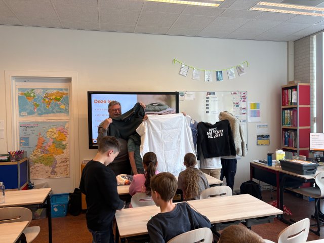 Wethouder van Drunen staat voor de klas met een shirt in zijn handen. Ook basisschoolleerlingen zijn zichtbaar. 