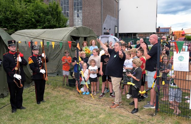 Wethouder Roland knipt juichend een lint door om de camping te openen. Op de achtergrond het Westhoes en een groep volwassenen en kinderen. Op de voorgrond twee mensen in uniform. 