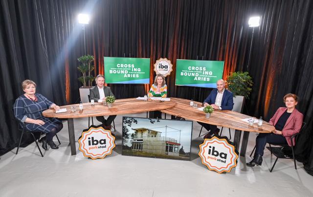 Het panel van de Closing Conference achter een tafel, logo van IBA op de voorgrond en t.v.-schermen op de achtergrond