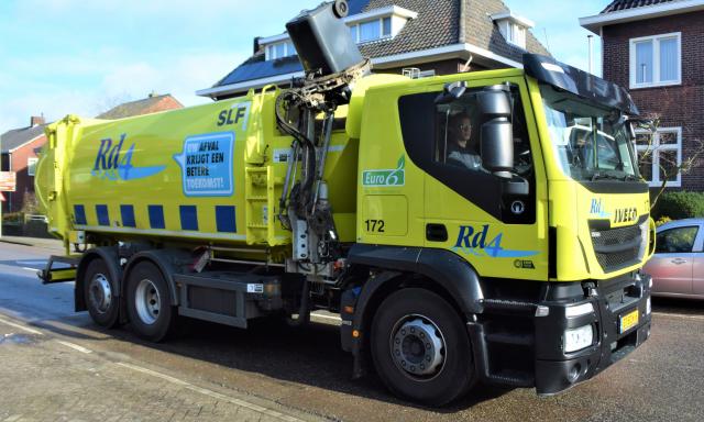 Een Rd4 vuilniswagen voor containers leegt op straat een container. Op de achtergrond zijn huizen te zien. 