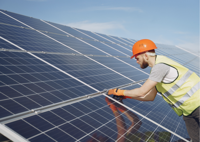 Een volwassene met een veiligheidsvest aan en een veiligheidshelm op controleert zonnepanelen op een dak.