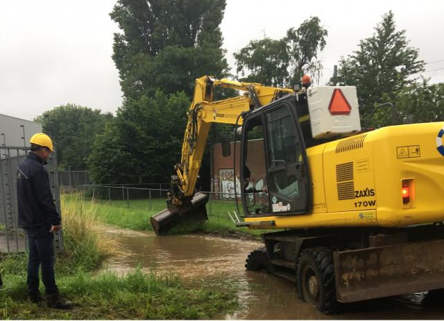 Een gele kraan graaft een gleuf in het gras op een plek waar het water hoog staat. Een persoon met een gele bouwhelm kijkt toe. 