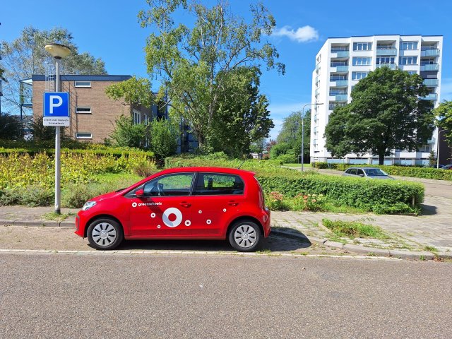 Een rode Volkswagen UP staat op een deelauto parkeerplaats aan de Kremerstraat. Op de achtergrond zie je gebouwen. 