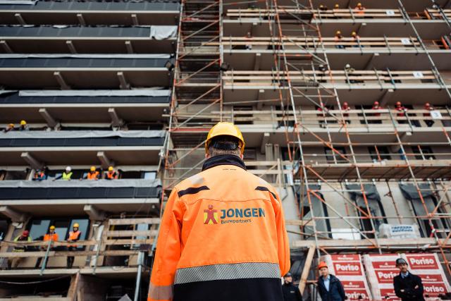 Bouwvakkers staan op de galerijen van de flat, zij worden toegesproken door iemand van wie we alleen de rug zien, deze persoon draagt een veiligheidshelm en  een veiligheidsjas van Jongen Bouwpartners