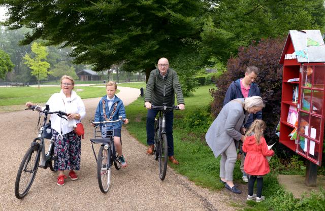 Van links naar rechts organisator Marleen Fischer, kinderburgemeester Hidde Sieler en wethouder Jo Schlangen op de fiets bij een minibieb waar een gezin een boek uitzoekt