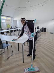 Fanta is in a conference room, holding a mop. She is also wiping down a tabletop with a cloth.