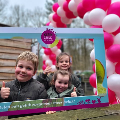 Kinderen staan achter een fotolijst met de tekst "Delen van geluk zorgt voor meer geluk!" 