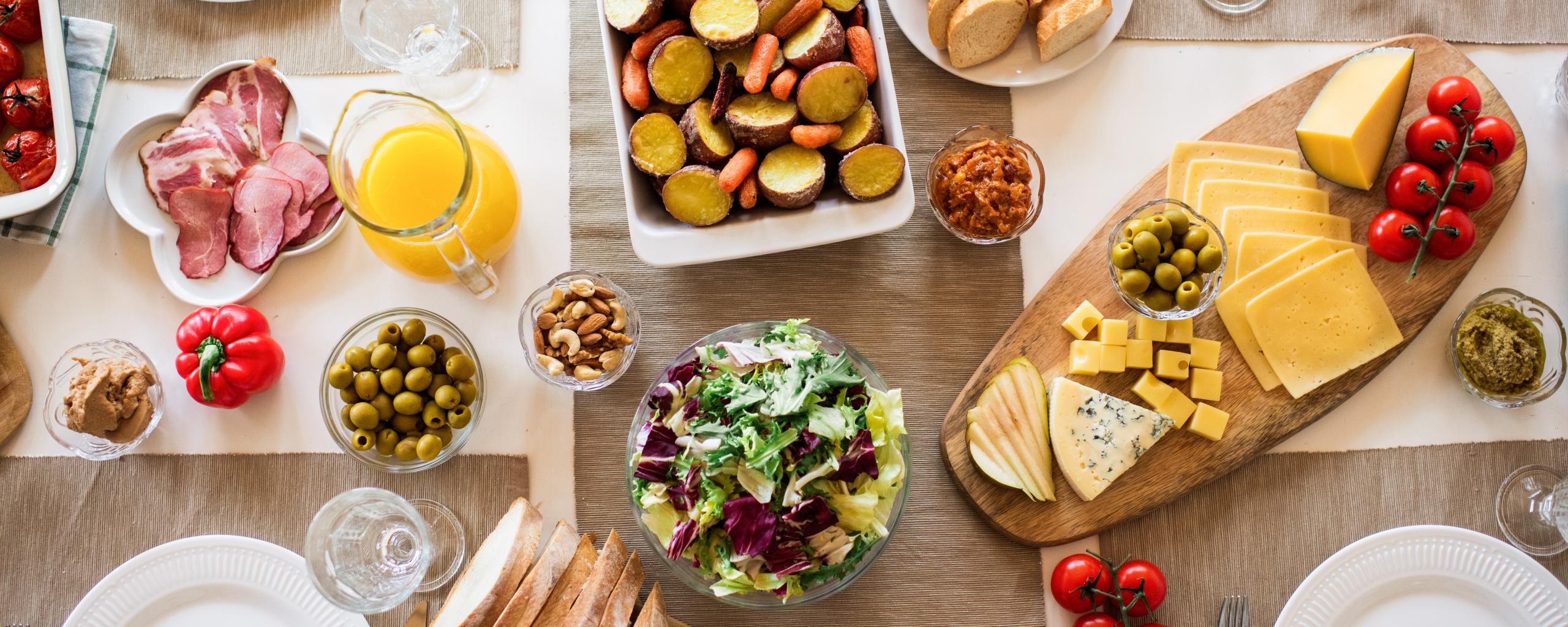 Bovenaanzicht van een gedekte tafel met onder andere een kaasplank, ovenschotel, diverse soorten vlees en olijven.