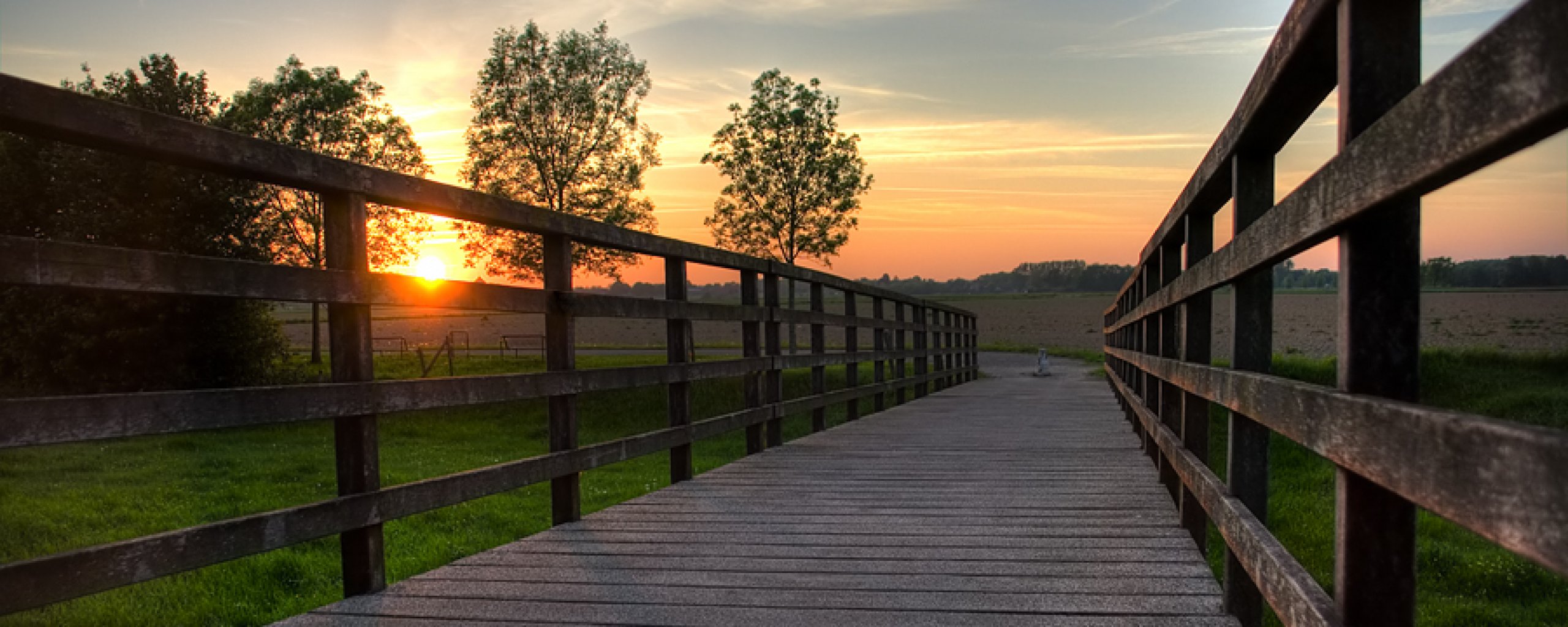 Een houten brug: de fietsbrug in Melick, met op de achtergrond gras, bomen en een zonsopkomst of zonsonderang.