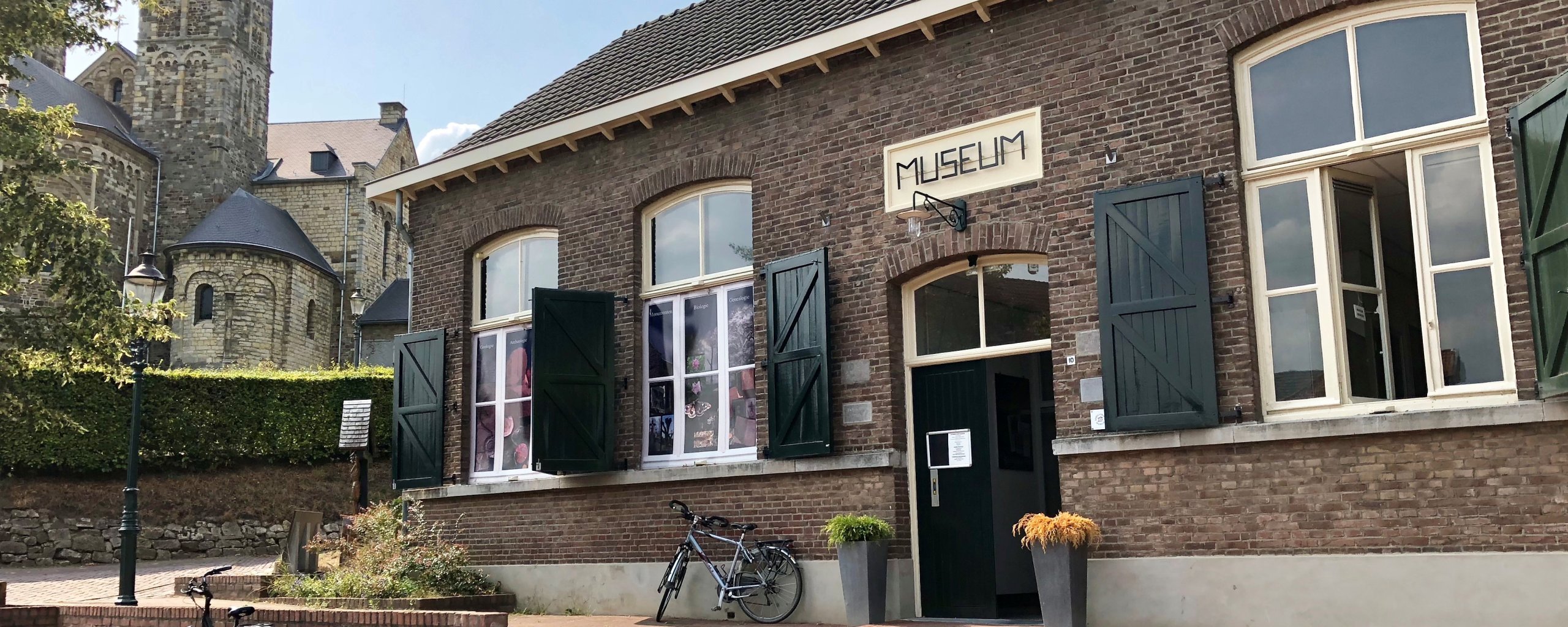 We zien een gebouw met groene luiken en een bord waar het woord 'museum' op staat geschreven. Dit is het Roerstreerkmuseum in Sint Odiliënberg. Op de achtergrond zien we één van de torens van de basiliek.