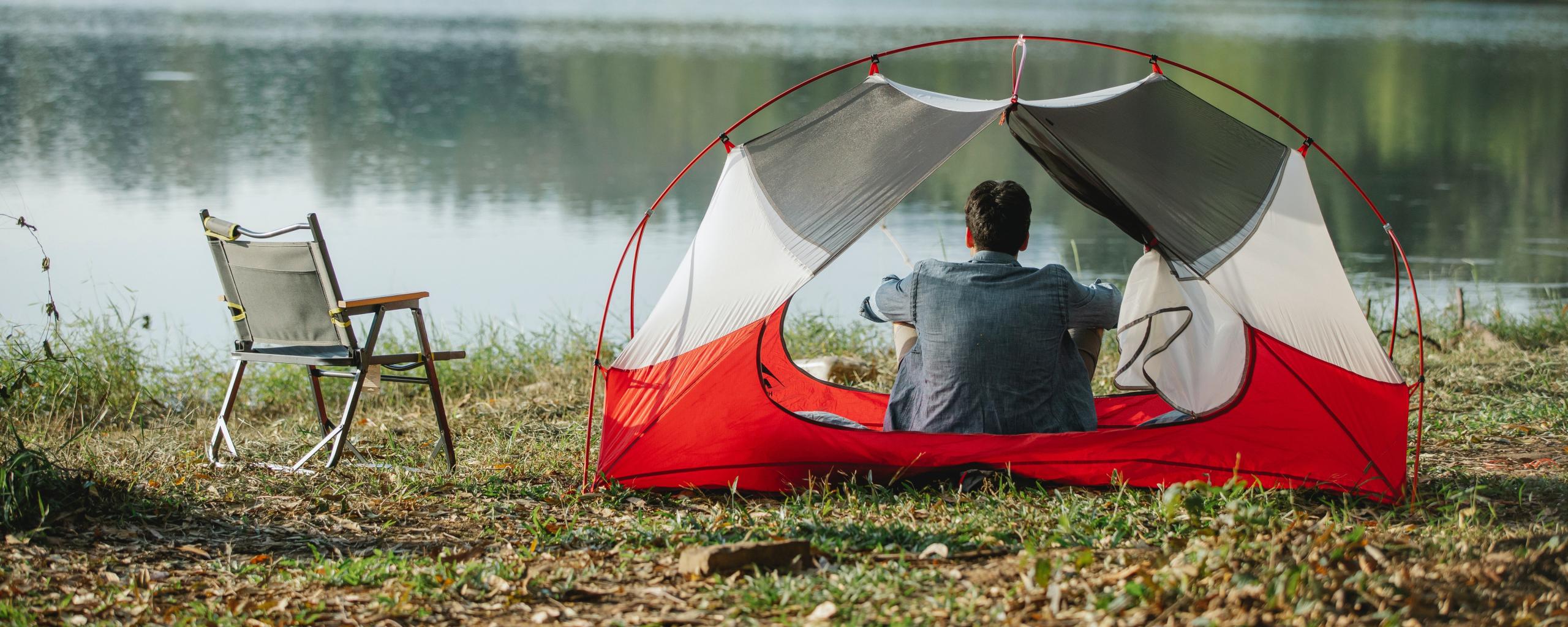 We zien iemand in een kampeertent zitten die uitkijkt over het water. Naast de tent staat een campingstoel.