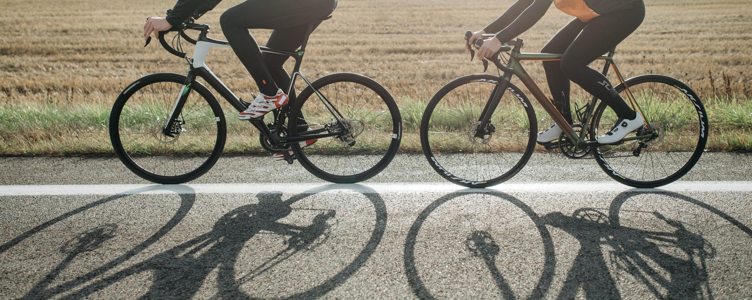 Twee mensen op een wielrenfiets. De mensen fietsen achter elkaar en we zien hen allebei van de zijkant. Ze fietsen op een weg en op de weg is de schaduw van de fietsen zichtbaar. Op de achtergrond zien we een velt.