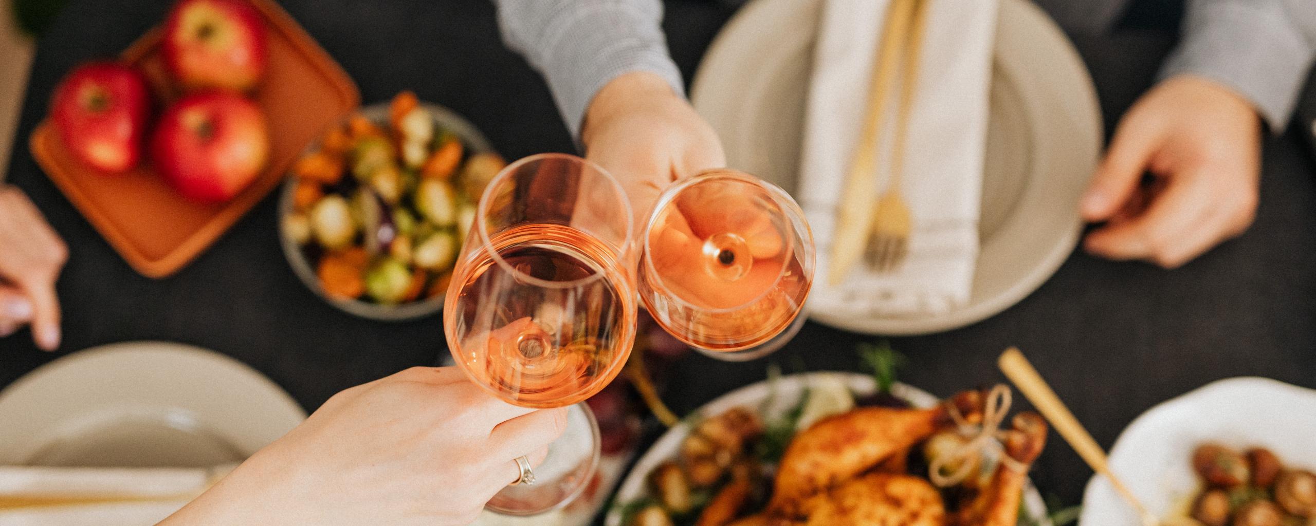Het bovenaanzicht van een tafel vol met eten. We zien twee handen die twee wijnglazen vasthouden en met elkaar proosten.