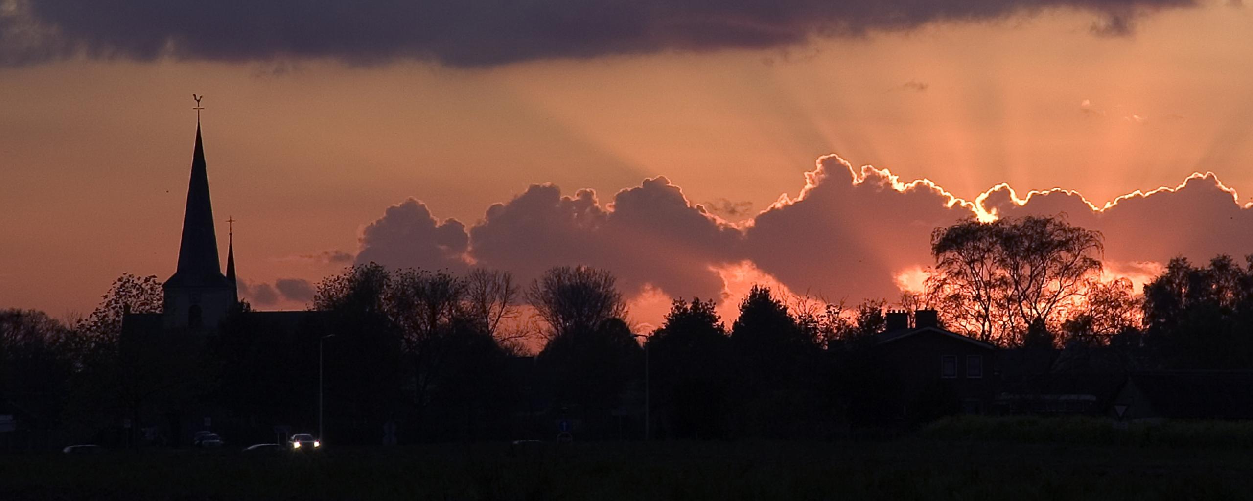 We zien de wolken en een oranje lucht waarin de zon ondergaat. Links in beeld is de kerktoren van Posterholt te zien.