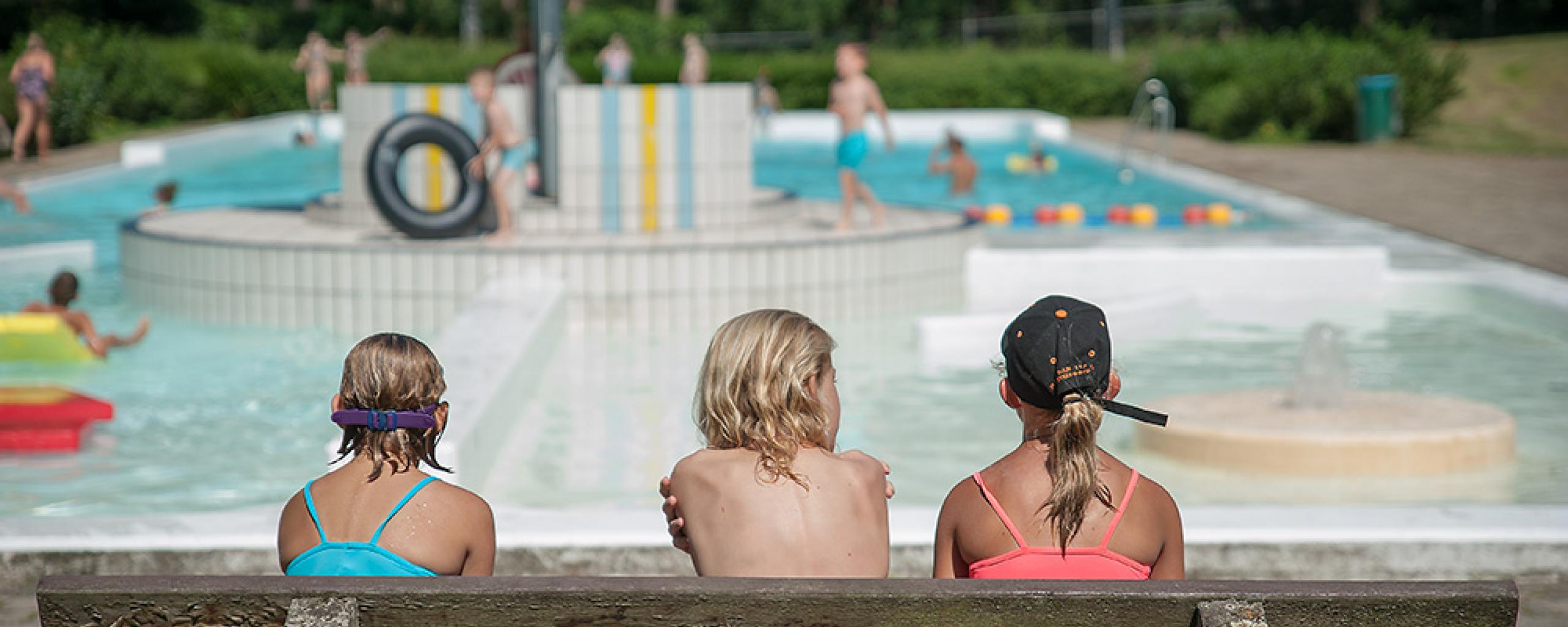Drie meisjes in zwemkleding op een bankje. We zien de meisjes van de achterkant. Ze kijken uit op zwembad 't Sweeltje.