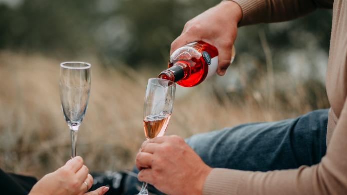 We zien twee glazen champagne die worden ingeschonken met een rood drankje. De mensen die het drankje inschenken liggen tussen het droge gras.