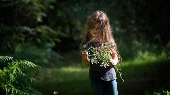 Een jong meisje met lange, bruine haren dat een bosje met witte bloemen vasthoudt. Het meisje kijkt naar de bloemen en staat tussen de planten en bomen van het Bergerbos.