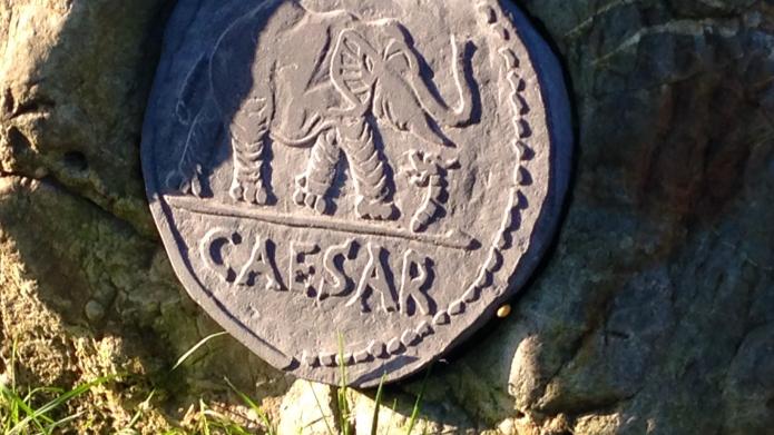 We zien een uitvergroot voorbeeld van een Romeinse munt. Op de munt staat een olifant afgebeeld en het woord 'Caesar'. De munt is bevestigd op een steen en is een voorbeeld van een munt die op deze geluksplek werd gevonden in 2009.