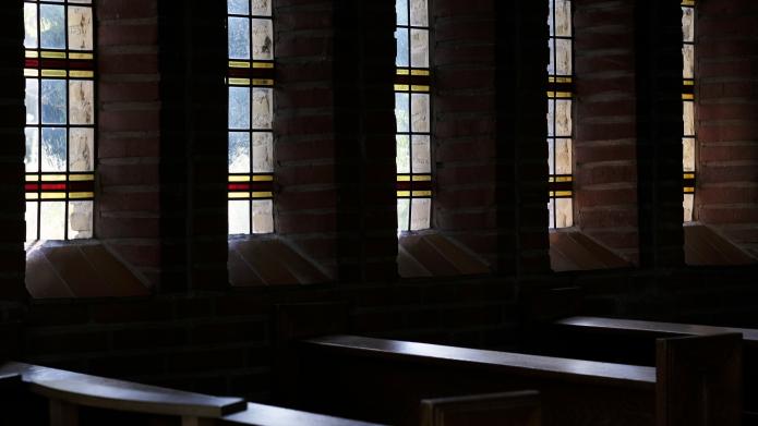 We zien de binnenkant van de kapel van de WIjngaard, een rij kerkbanken met op de achtergrond geruite glas in lood ramen die daglicht naar binnen laten schijnen.