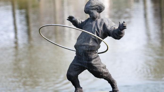 Een standbeeld op een sokkel in het water. Het standbeeld is een kind dat speelt met een hoepel.