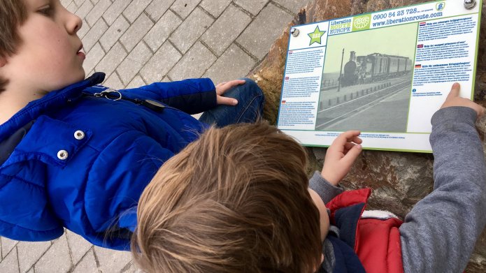 Twee kinderen die naar een bordje kijken met informatie over een trein uit de tweede wereldoorlog. Het bordje is onderdeel van de Liberation Route.
