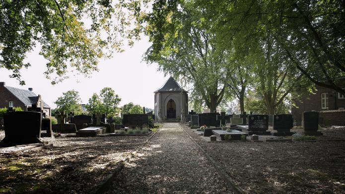 Een stenen pad met links en rechts grafstenen. Aan het einde van het pad is een kapelletje zichtbaar. Op het kerkhof valt een schaduw van hoge bomen.