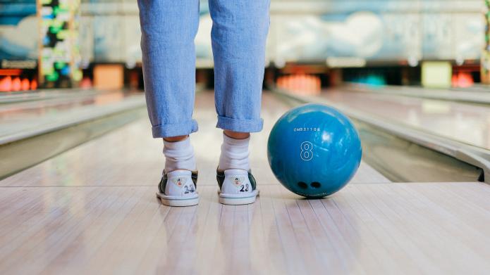 We zien de achterkant van de schoenen en broek van iemand die klaar staat om een bowlingbal te gooien op een bowlingbaan. We zien de glimmende vloer van de baan en naast de voeten ligt een blauwe bowlingbal.