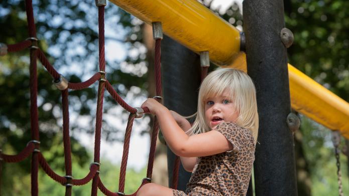 We zien een jong kind met blonde haren dat in een klimrek speelt. Het kind draagt een korte broek en een top met korte mouwen met een luipaardmotief.