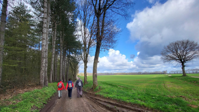 Wandelaars lopen langs de bosrand. Rechts een veld met een mooie blauwe lucht met witte wolken.