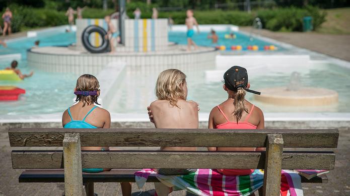 Drie meisjes in zwemkleding op een bankje. We zien de meisjes van de achterkant. Ze kijken uit op zwembad 't Sweeltje.