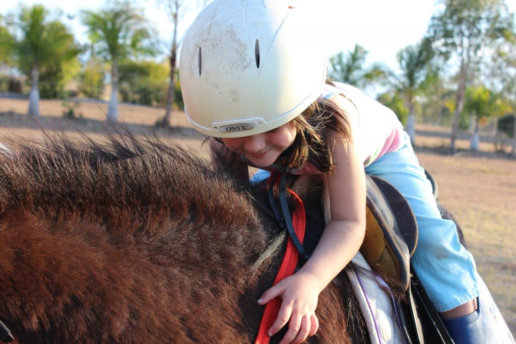 We zien een jong meisje met een witte helm op de rug van een paard. Het meisje lacht, buigt naar voren en houdt zich vast aan het paard.