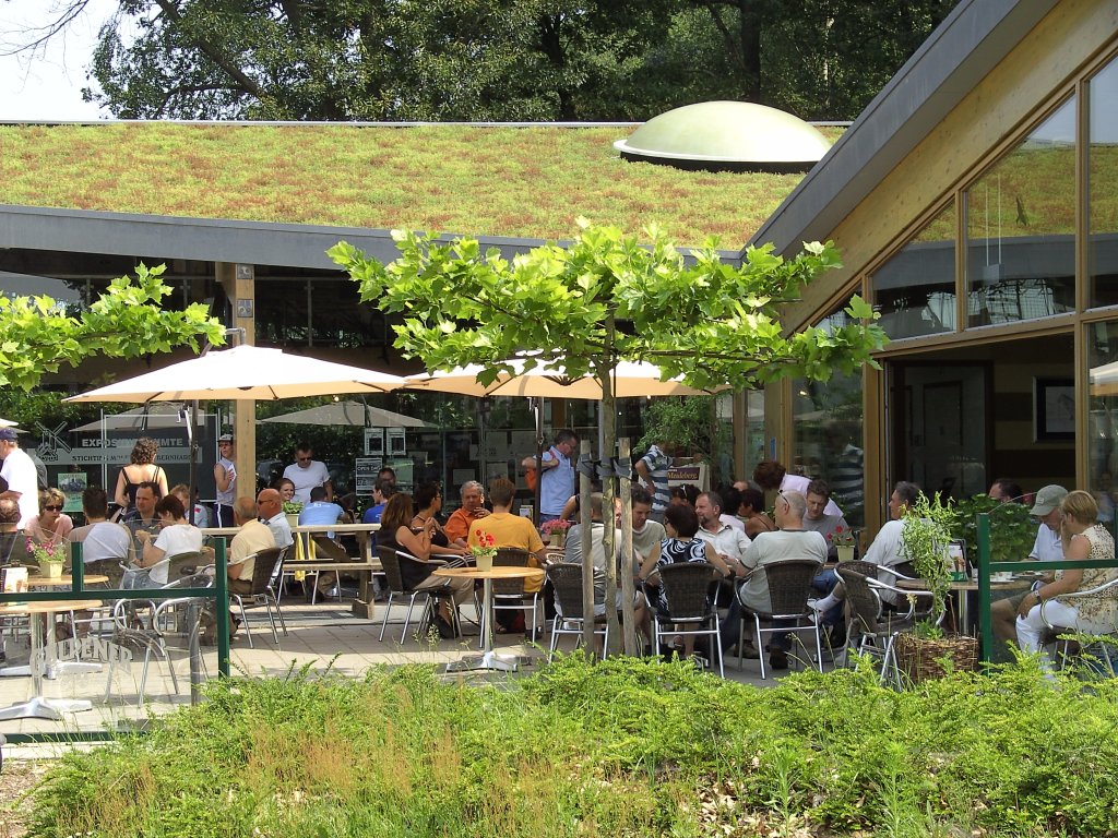 Gezellig ogend terras voor een gebouw met een groen dak en hoge ramen. Het terras heeft diverse tafeltje die allemaal vol zitten en diverse parasols. Er staat een bloembak met groene planten op de voorgrond, een er staan een paar leiboompjes op het terras.