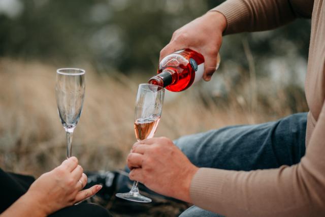 We zien twee glazen champagne die worden ingeschonken met een rood drankje. De mensen die het drankje inschenken liggen tussen het droge gras.