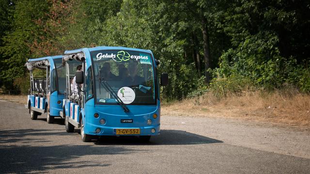 We zien een elektrisch shuttletrein met twee wagens: de Geluksexpres. De treintjes zijn blauw van kleur en rijden op een verharde weg met bomen aan de zijjkant van de weg.