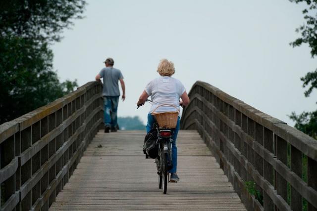 Een houten brug: de fietsersbrug in Melick. Op de brug fietst een mevrouw met een mandje achterop de fiets. Links van haar wandelt een meneer. We zien beiden personen van de achterkant de brug oversteken.