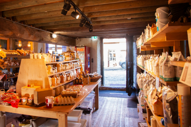 We zijn een winkel vol houten schappen met een houten vloer en houten plafondbalken. In de schappen staan onder andere brood, eieren, meel en jam.