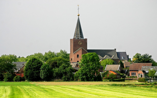 Zicht op de kerk in Herkenbosch. Op de voorgrond veld en bomen.