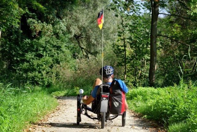 We zien een bospad op een zonnige dag met links en rechts veel groene planten. Op het pad zien we de achterkant van iemand op een ligfiets. Hij draagt een helm en een blauw t-shirt. Er hangen rode fietstassen op de ligfiets en achterop de fiets wappert een vlag met zwart, rood en geel: de Duitse vlag.