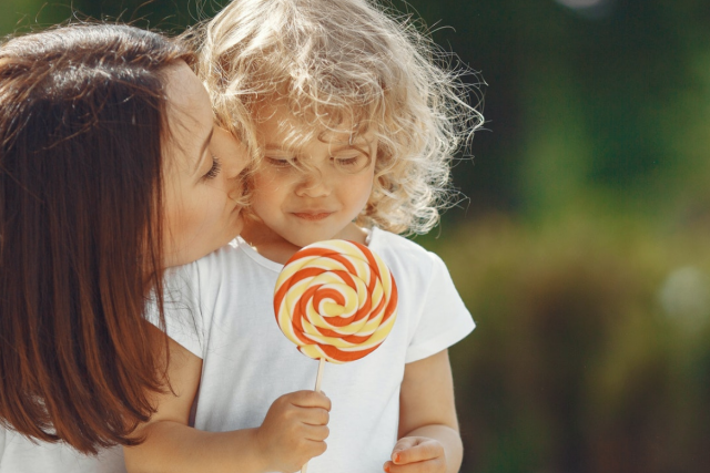 We zien een kind met blonde krullen dat een grote lolly vasthoudt. We zien links de moeder die het kindje een zoen op de wang geeft.