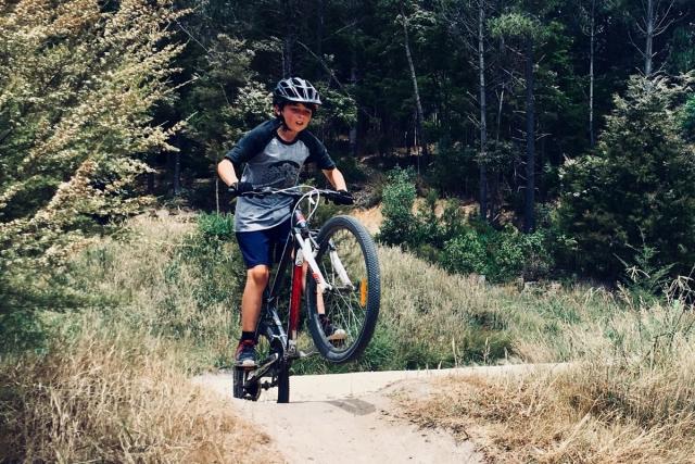 Een jonge jongen op een mountainbike op een pad tussen droog gras. Op de achtergrond zien we bomen.