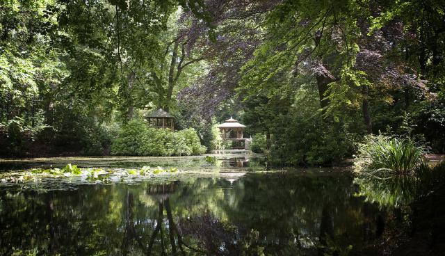 Een vijver omringt door hoge bomen. In de verte zien we twee prieeltjes. Links een houten huisje en rechts een houten prieeltje in Indiase bouwstijl: het paviljoen van het bospark.