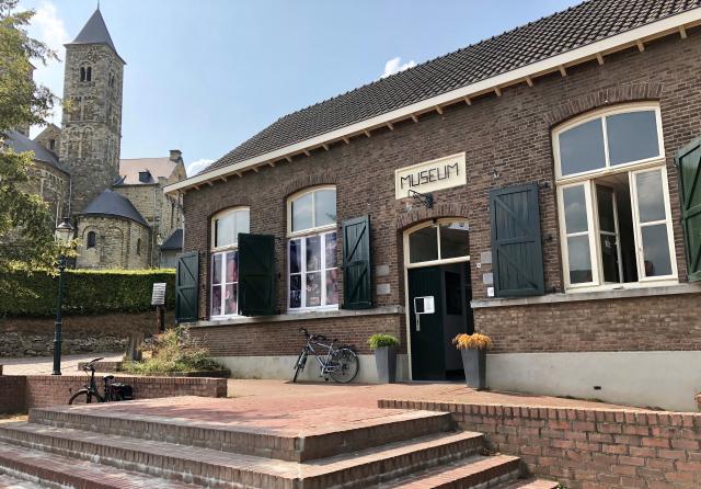 We zien een gebouw met groene luiken en een bord waar het woord 'museum' op staat geschreven. Dit is het Roerstreerkmuseum in Sint Odiliënberg. Op de achtergrond zien we één van de torens van de basiliek.