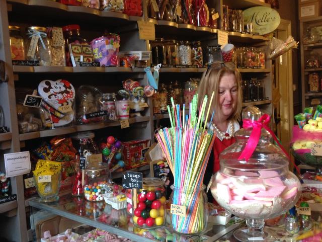 We zien Marie-Anne, de eigenaresse van het snoepwinkeltje in Posterholt achter de toonbank van haar winkel, omringt door potten met gekleurde snoepjes.