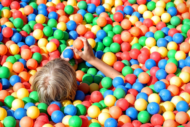 We zien een ballenbak vol gekleurde balletjes. In de ballenbak zien we de achterkant van het hoofd van een kind. De rest van het lijf is verstop in de ballenbak.