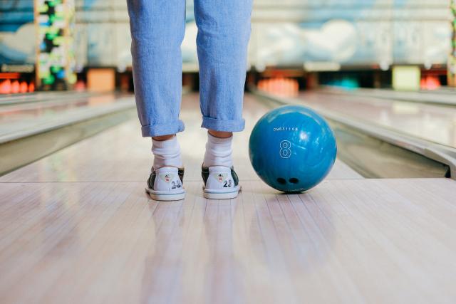 We zien de achterkant van de schoenen en broek van iemand die klaar staat om een bowlingbal te gooien op een bowlingbaan. We zien de glimmende vloer van de baan en naast de voeten ligt een blauwe bowlingbal.